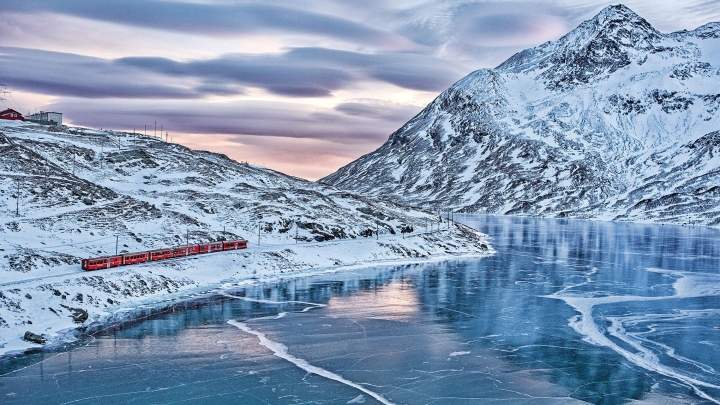 The Bernina Express train next to the Lago Bianco covered in black ice in winter.